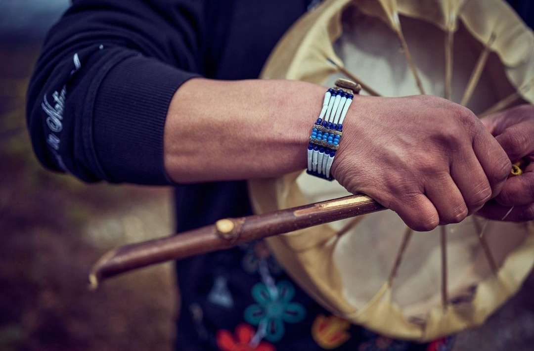 Close-up of a person holding a drum and drumstick, wearing a beaded bracelet, in a blurry, natural outdoor setting.