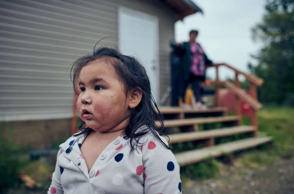 Portrait of a young girl with a polka dot shirt, standing outside near a house with an adult in the background on the porch.