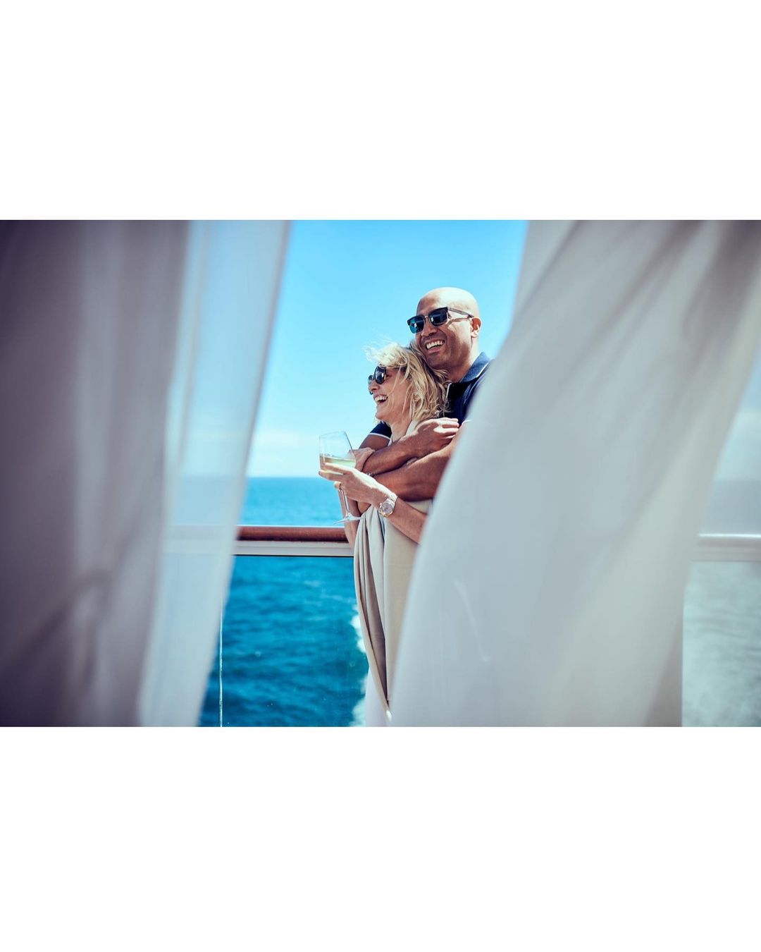 Couple enjoying drinks on a cruise ship deck with blue ocean and sky, framed by white curtains, creating a romantic travel image.