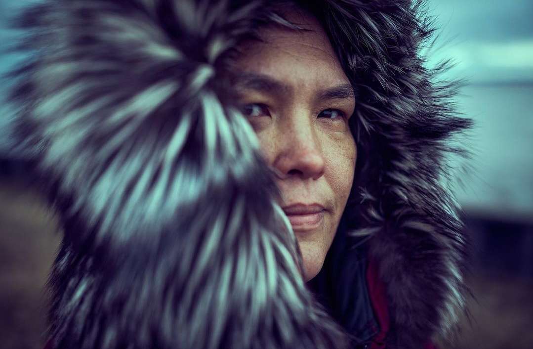Close-up portrait of an adult with a neutral expression wearing a fur-lined hood, set against a blurry outdoor background.