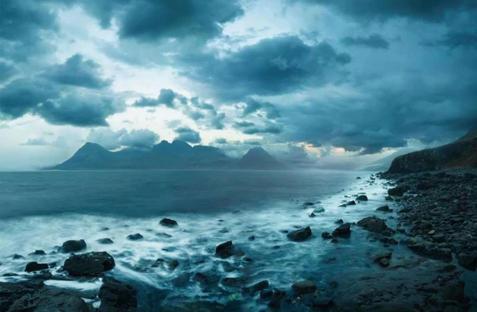 Scenic seascape view featuring a rocky shoreline, the ocean, and distant mountains under a dramatic, cloudy sky with a blue hue.