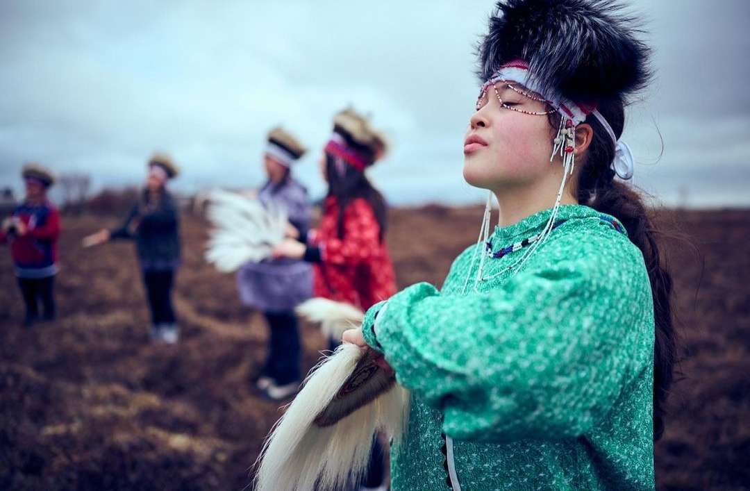 Group of Inuit women in traditional dress performing outdoors with white feathered fans against a cloudy sky in a natural tundra environment.