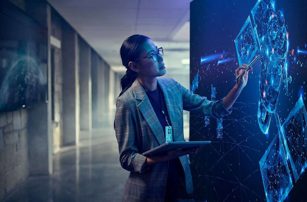 Adult female in business attire interacts with a digital screen while holding a tablet in a modern office hallway setting.
