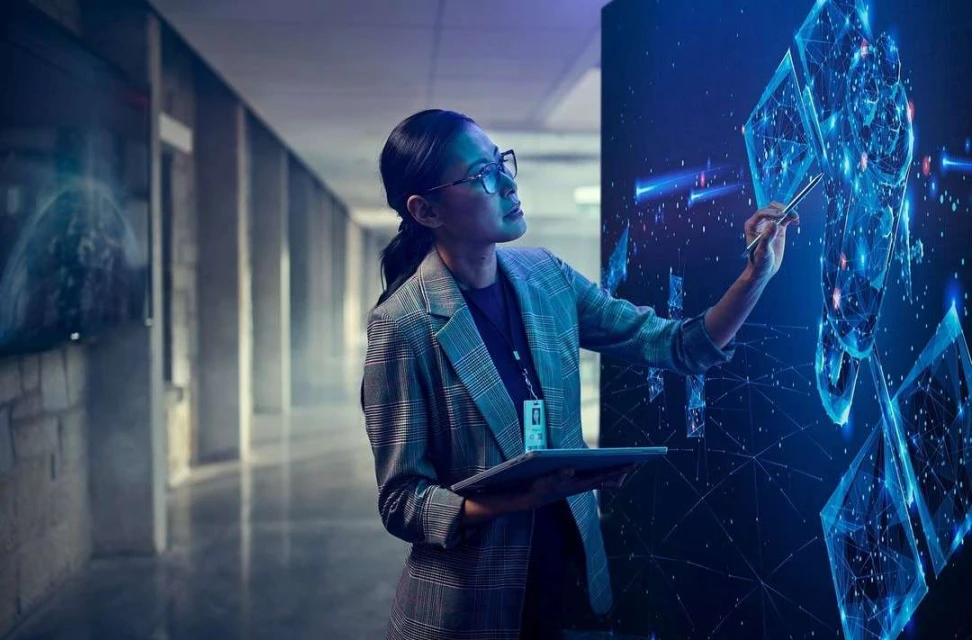 Adult female in business attire interacts with a digital screen while holding a tablet in a modern office hallway setting.