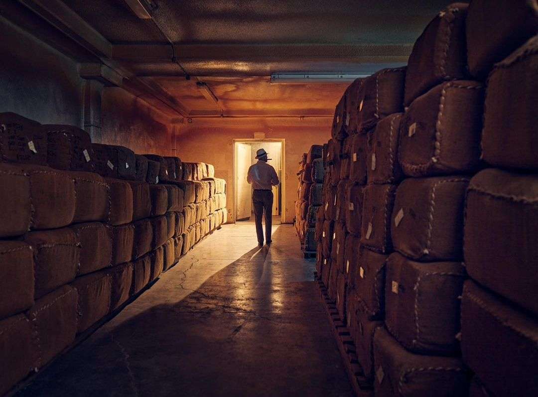 Warehouse interior with a man walking toward a doorway, surrounded by stacks of burlap sacks, with low, warm lighting.