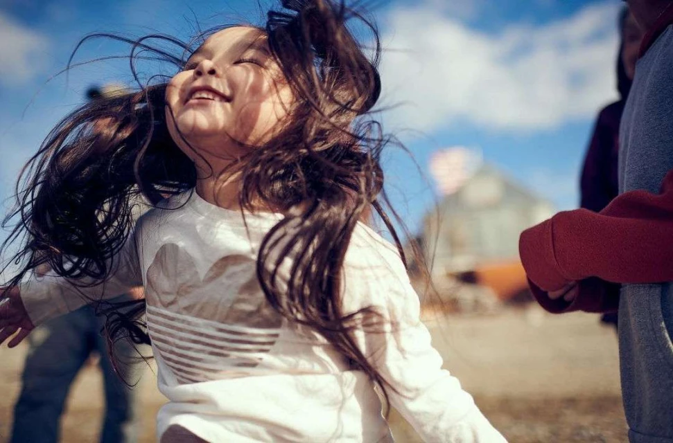 Happy child with long hair leaps outdoors against a blue sky in a white long-sleeved shirt, with other figures blurred in the background.