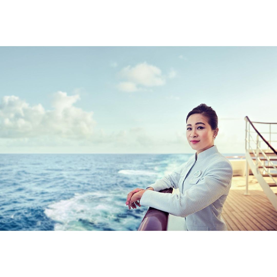 Woman in jacket stands at the railing of a cruise ship at sea. Blue sky, blue water, and the ship's deck fill the background.