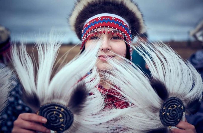 Portrait of a young woman in traditional Indigenous fur clothing and headdress holding feathered objects outside on an overcast day.