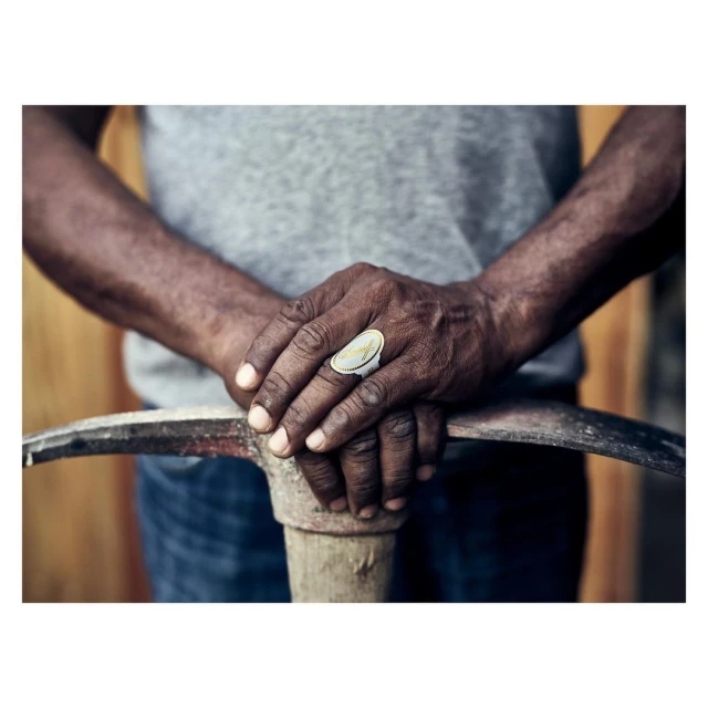 Close-up of a dark-skinned adult male's hands with a ring, resting on a pickaxe, wearing a grey shirt against a rustic wooden background.