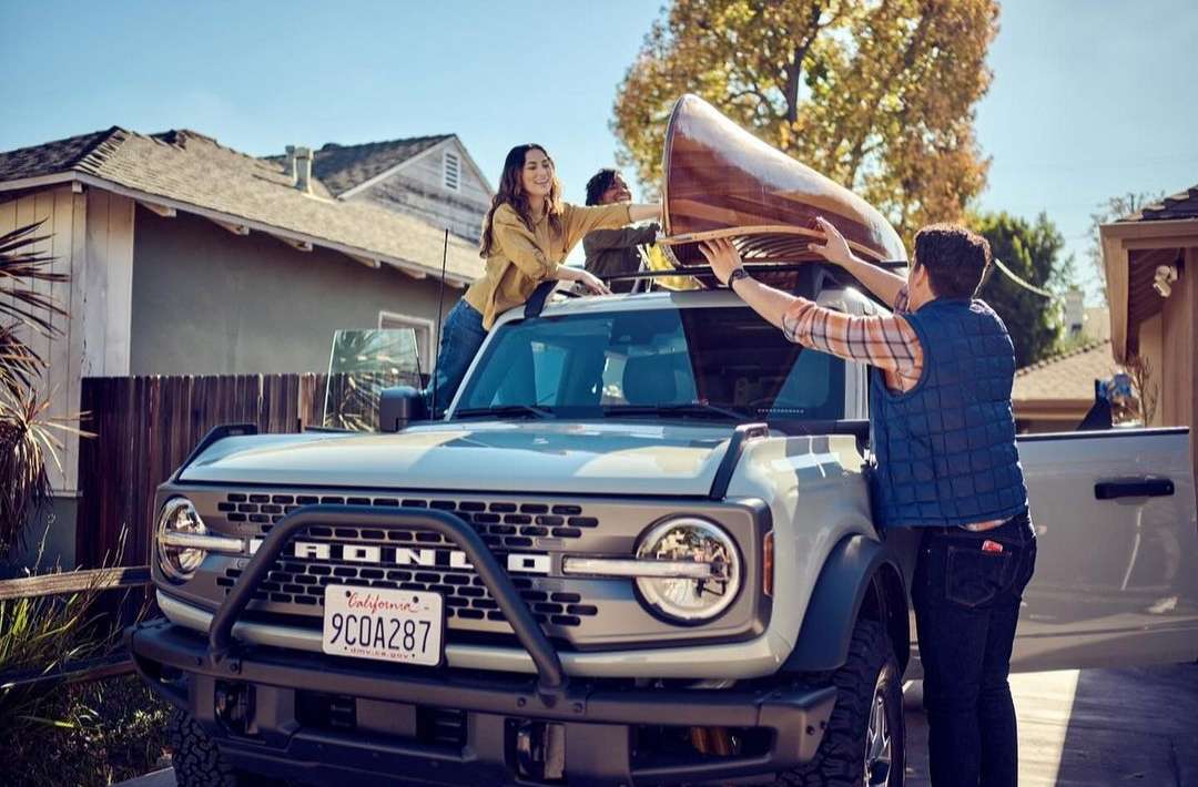 Ford Bronco SUV parked on a residential street as three people load a canoe on the roof rack against a clear blue sky.