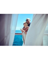 An older couple in sunglasses embracing on a cruise ship balcony overlooking the ocean, framed by white curtains.