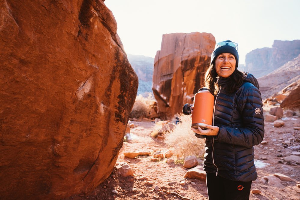 A woman in a black puffer jacket holds an orange water bottle in a desert landscape with large rock formations on a sunny day.