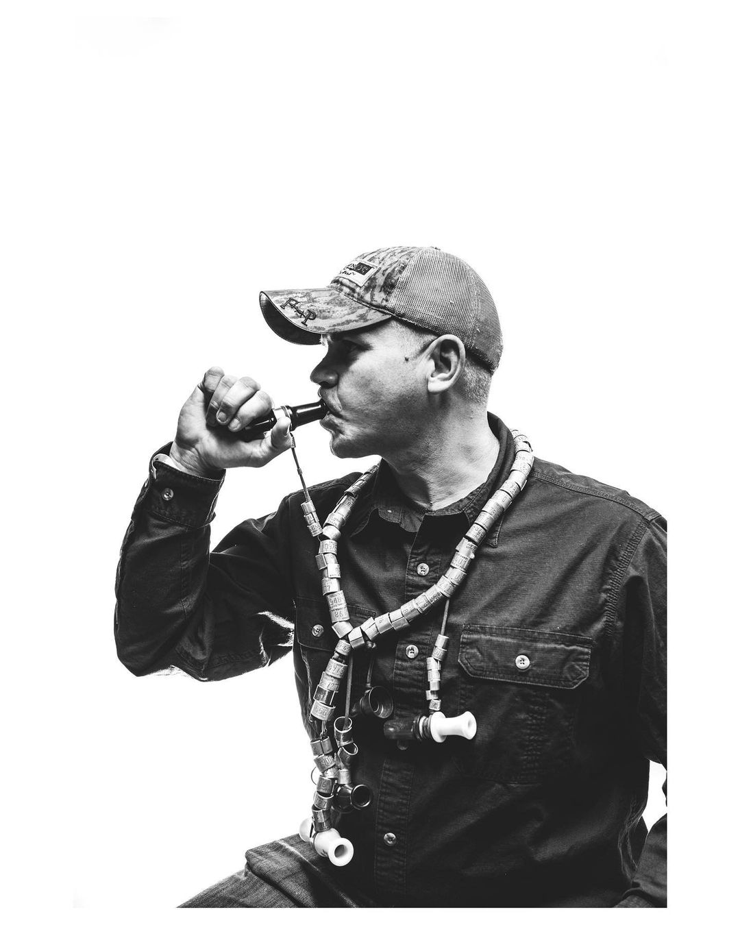Monochrome close-up of a man in a camo hat blowing a duck call while wearing a lanyard of calls against a bright white background.