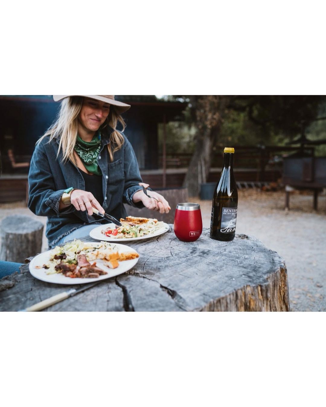 Woman in hat eats outdoors at stump table with drinkware and bottle. Plates of food feature grilled meats and salad, rustic background.