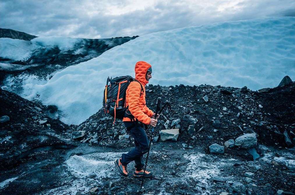 A man hikes across a rocky, icy landscape with a backpack and trekking poles, wearing an orange jacket, under a cloudy sky.