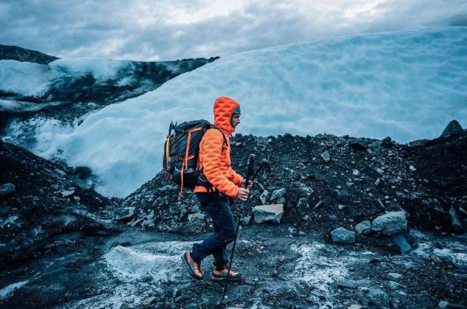 A man hikes across a rocky, icy landscape with a backpack and trekking poles, wearing an orange jacket, under a cloudy sky.