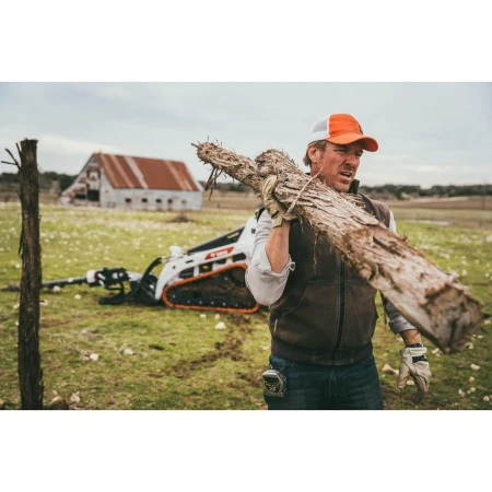 Man carries weathered wood on shoulder in rural field with compact tractor and barn, under overcast sky, wearing cap and work clothes.