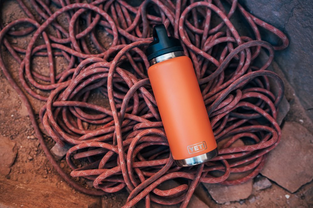 An orange Yeti water bottle sits on a pile of coiled red and black climbing rope, set against a red-rock textured surface.