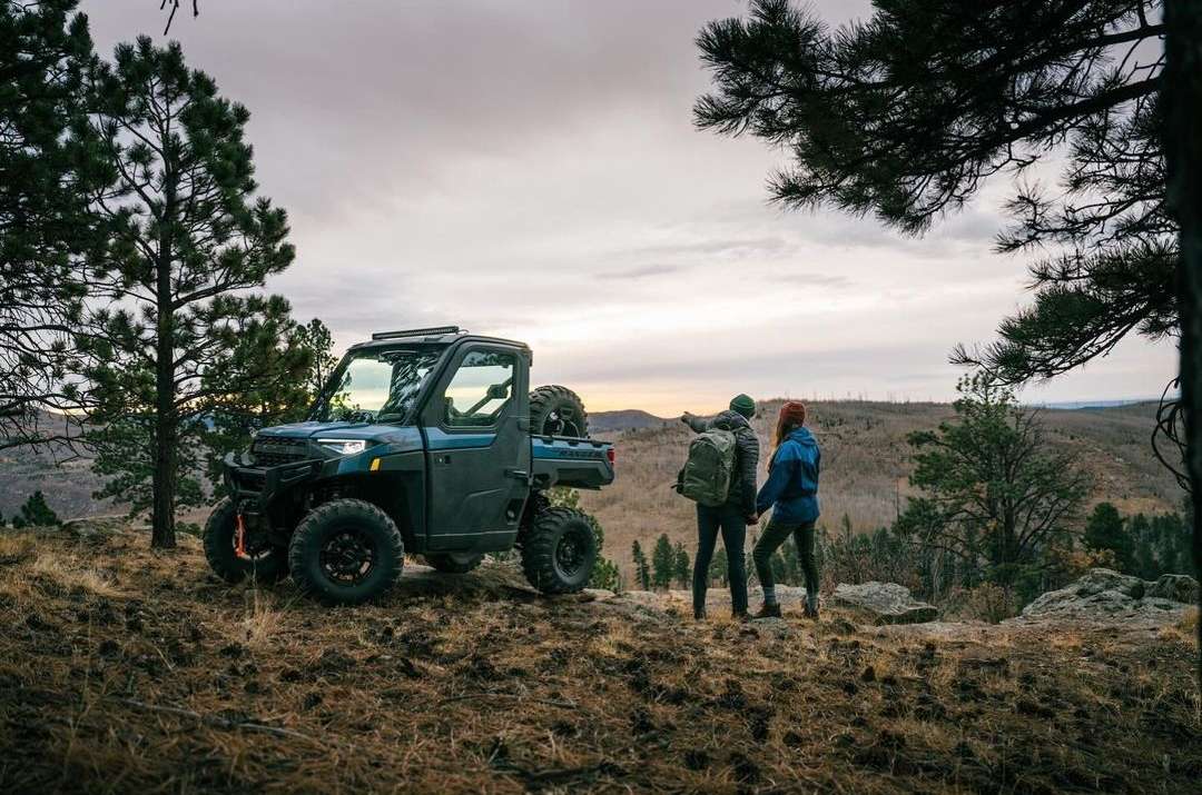 Off-road vehicle parked on a hillside with two people looking at the landscape, featuring trees and a cloudy sky at dusk.