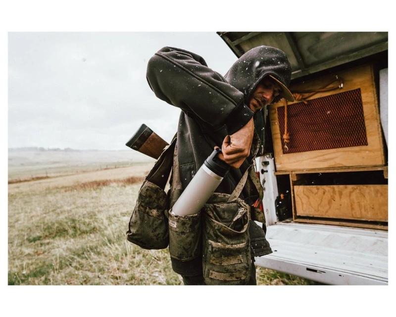 Man in hooded jacket standing next to pickup truck holding silver tumbler with grassy field and cloudy sky in the background.