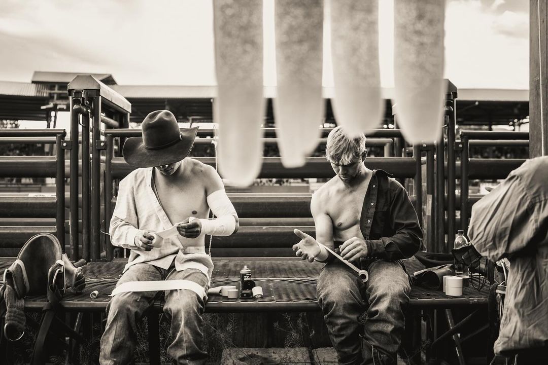 Black and white shot of two shirtless cowboys sitting outdoors with gear, one wrapping his arm, the other holding a tool.
