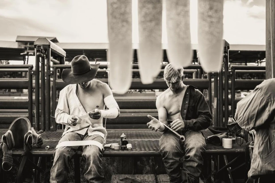 Black and white shot of two shirtless cowboys sitting outdoors with gear, one wrapping his arm, the other holding a tool.