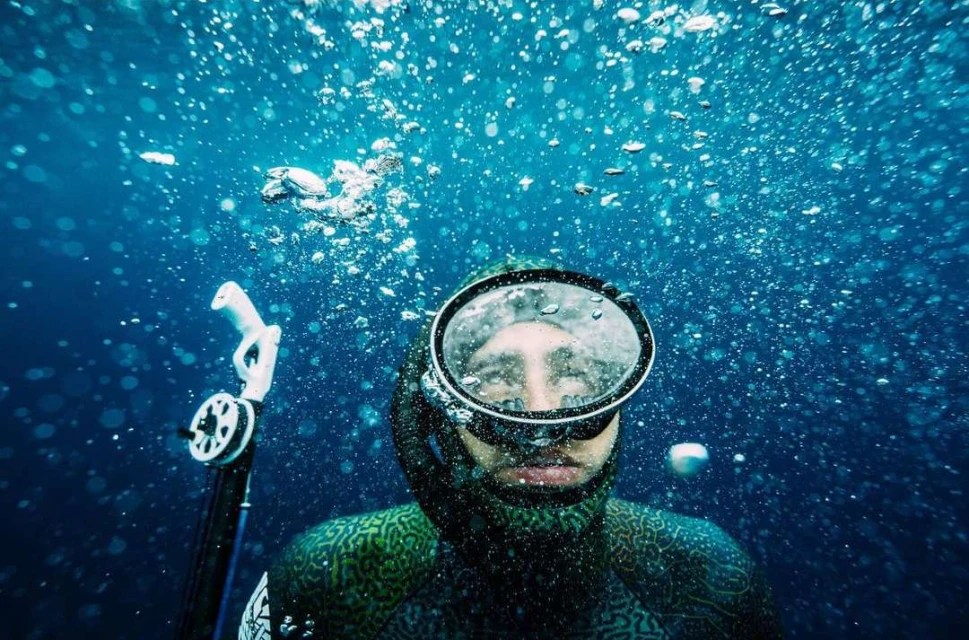 Underwater view of a free diver in a wetsuit and mask, bubbles rising toward the light blue water surface.