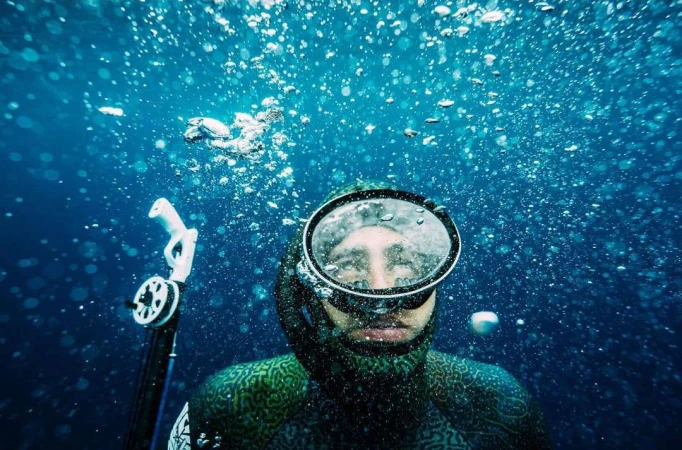 Underwater view of a free diver in a wetsuit and mask, bubbles rising toward the light blue water surface.