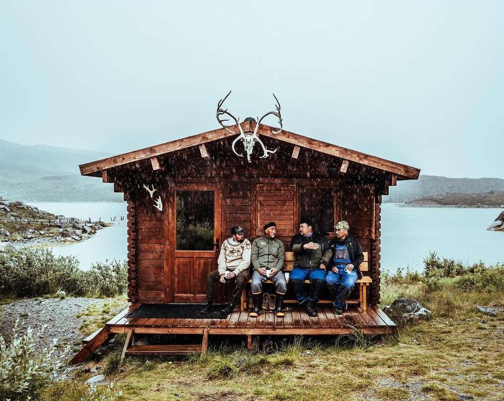 Four men sit on a porch of a log cabin on a rainy day in a mountainous landscape with a lake and vegetation in the background.