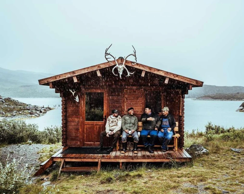 Four men sit on a porch of a log cabin on a rainy day in a mountainous landscape with a lake and vegetation in the background.