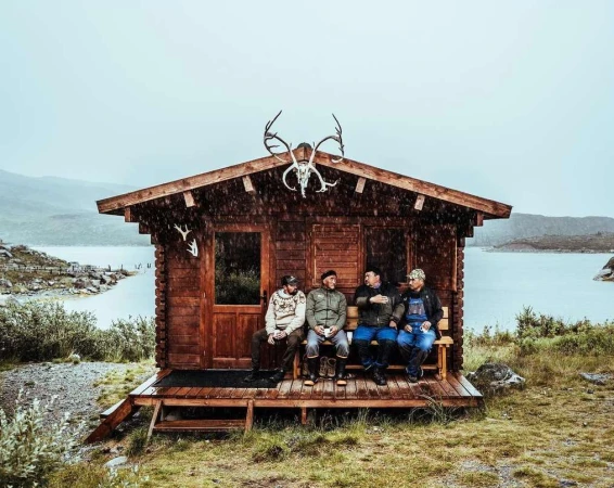 Four men sit on a porch of a log cabin on a rainy day in a mountainous landscape with a lake and vegetation in the background.