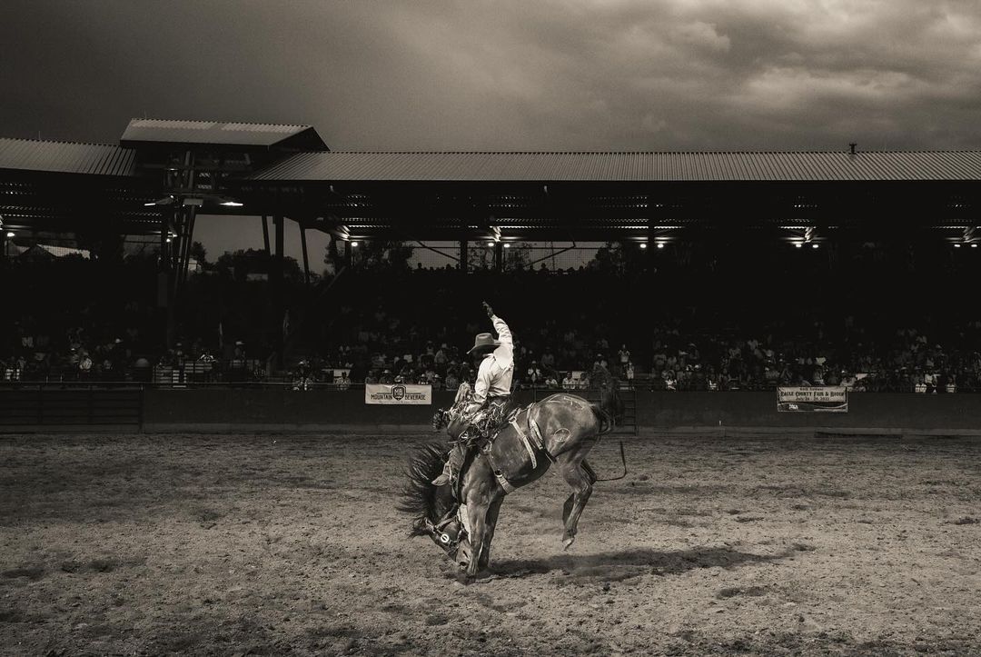 Monochrome photo of a rodeo cowboy riding a bucking bronco in an arena, with spectators in the background, under a dramatic sky.