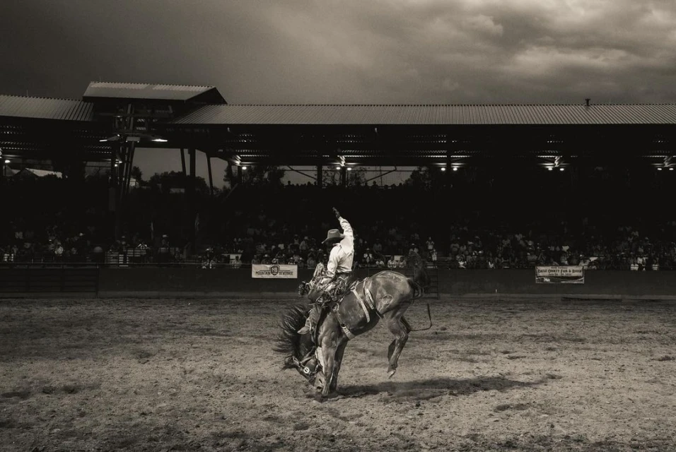 Monochrome photo of a rodeo cowboy riding a bucking bronco in an arena, with spectators in the background, under a dramatic sky.
