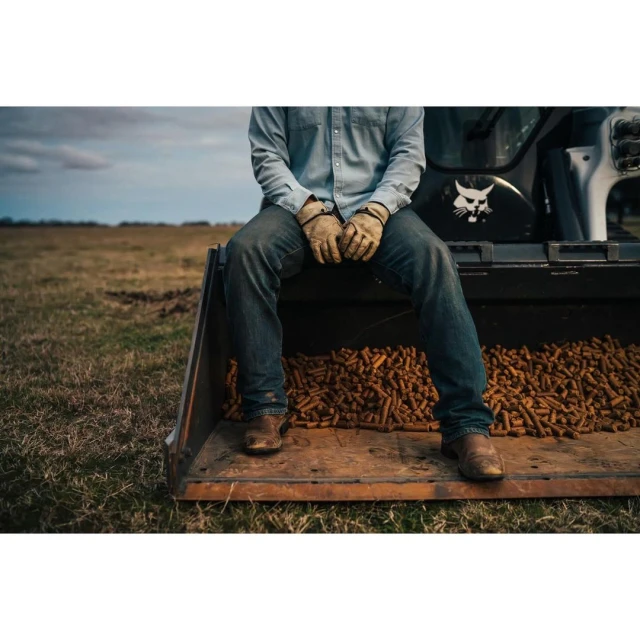 Person sits in the bucket of a construction loader filled with cylindrical food pellets outdoors on a farm, wearing jeans and denim shirt.