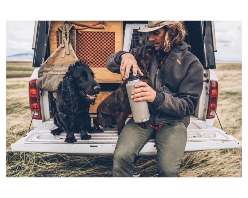 Man sitting on pickup truck tailgate with two dogs in a grassy field. He is opening a water bottle while outdoors.