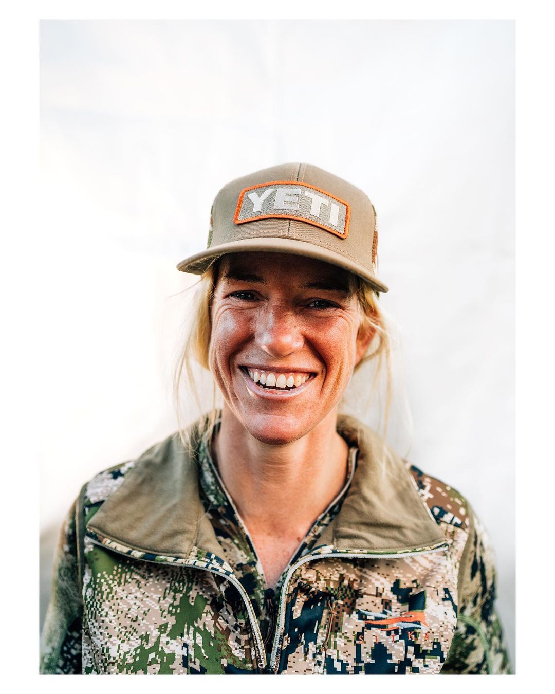A woman smiles wearing a tan Yeti baseball cap and camo jacket, showcasing outdoor and active lifestyle themes with a bright background.