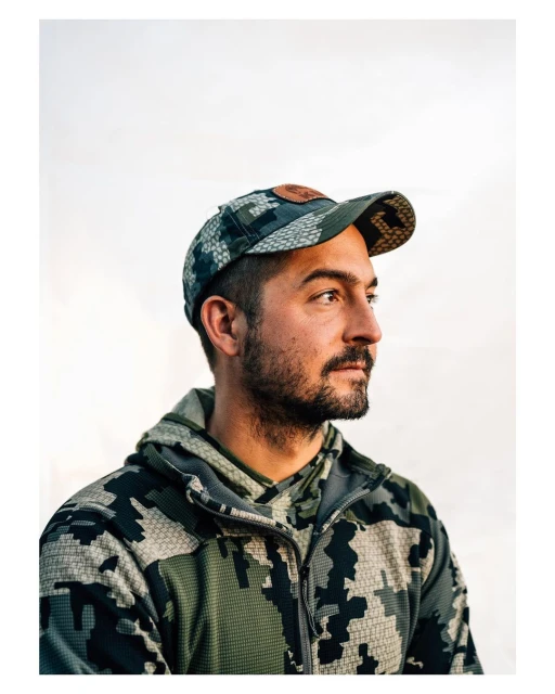 Portrait of a man with a beard wearing a camouflage baseball cap and hooded jacket against a white backdrop, looking off to the side.