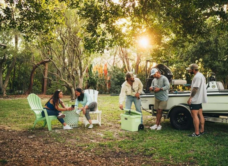 Group of five people gathered near a boat and a cooler in a grassy, tree-filled outdoor setting, enjoying a sunny day.