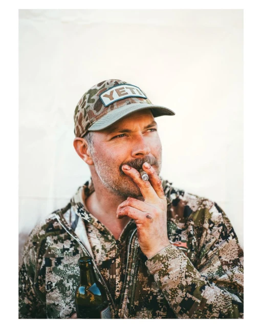 Adult male in camo hat and jacket smokes a cigar and holds a beer bottle against a white background in a medium studio shot.