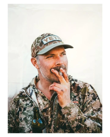 Adult male in camo hat and jacket smokes a cigar and holds a beer bottle against a white background in a medium studio shot.