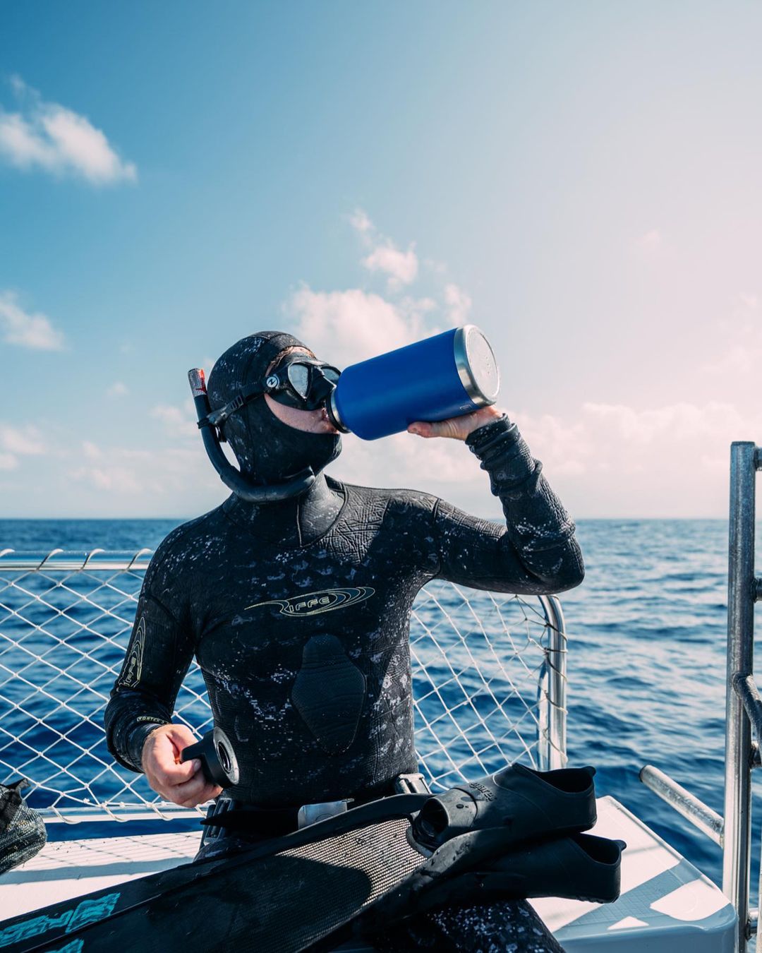 Person in wetsuit drinking from a blue water bottle on a boat with ocean and sky background, sunny day.