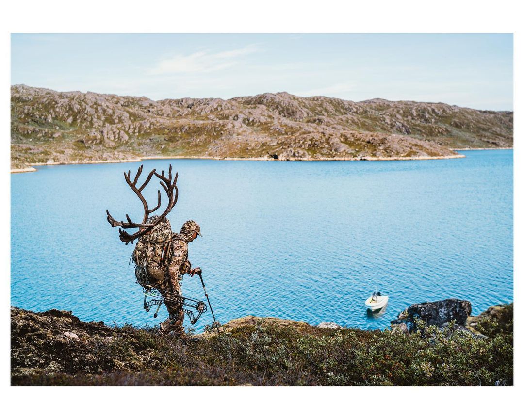 Hunter treks through rocky terrain alongside blue water with a large set of antlers strapped to his back, carrying a bow and walking stick.