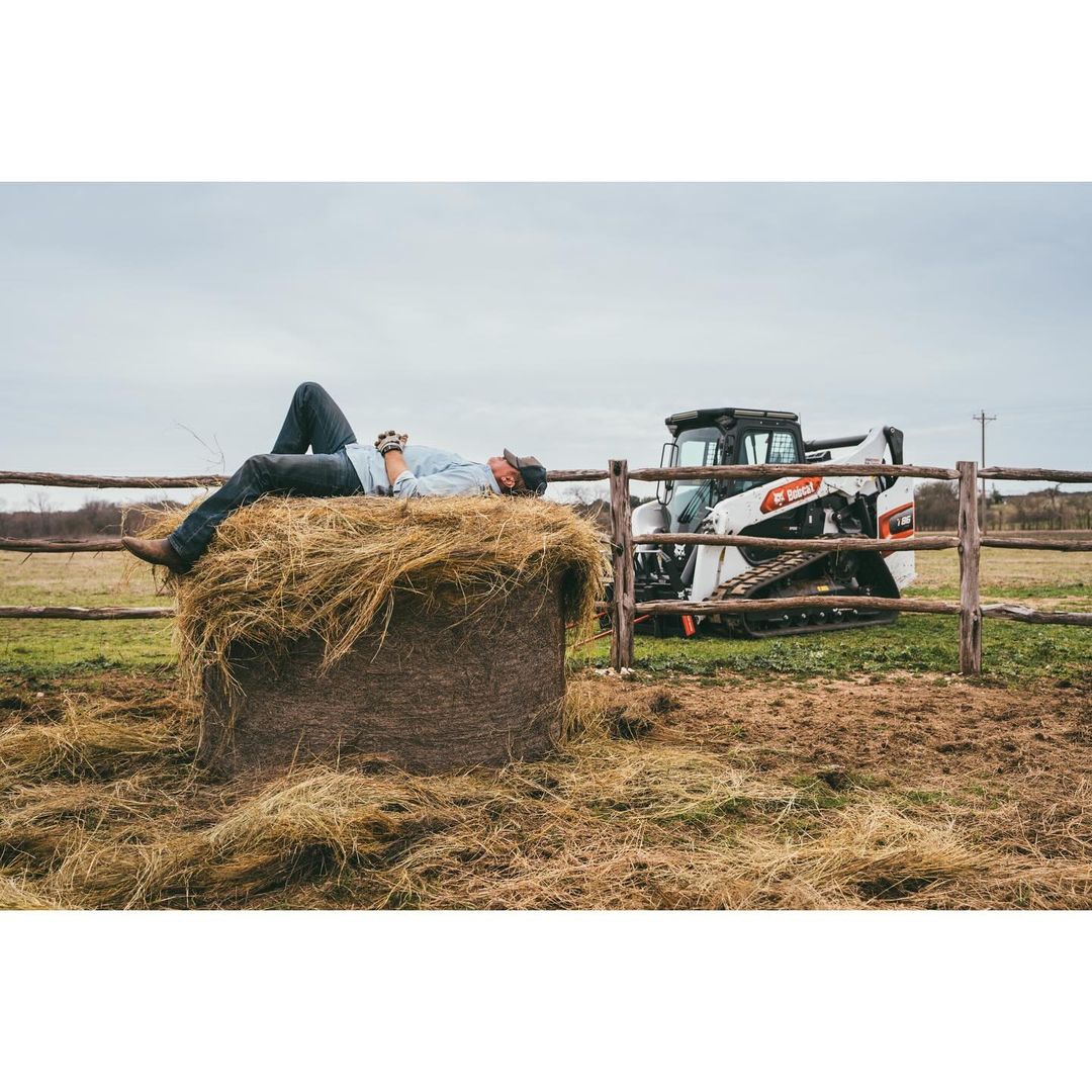 Adult male relaxing on a hay bale with a Bobcat skid-steer loader in the background, taken outdoors on a farm under a cloudy sky.