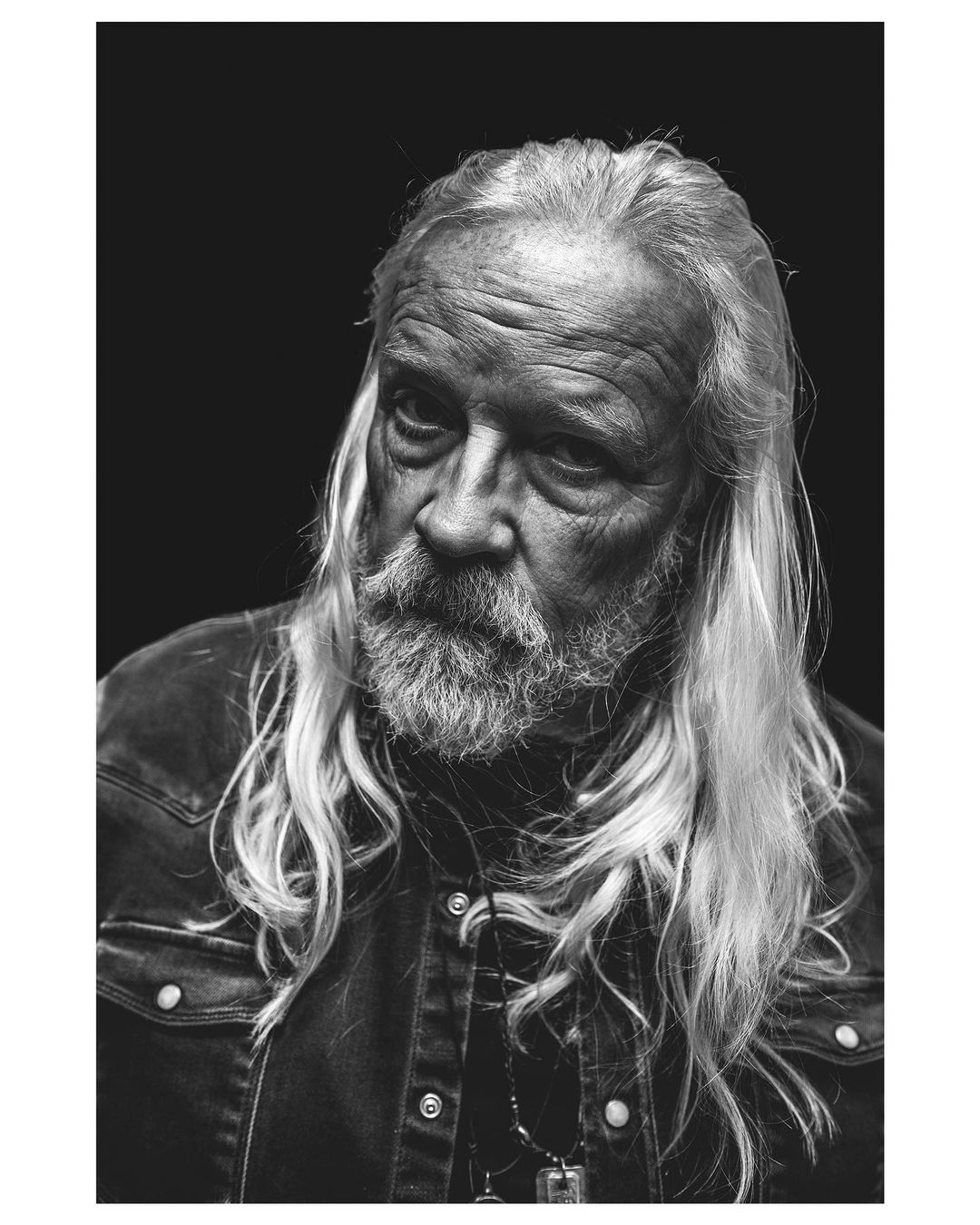 Monochrome portrait of an elderly man with long white hair and a beard, wearing a denim jacket against a dark backdrop.