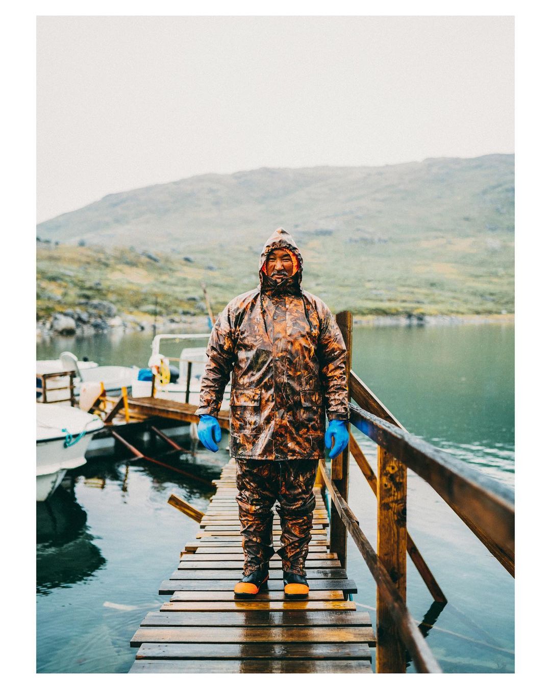 Fisherman in full waterproof gear stands on a wooden pier with boats and a green, hilly landscape visible beyond the lake.