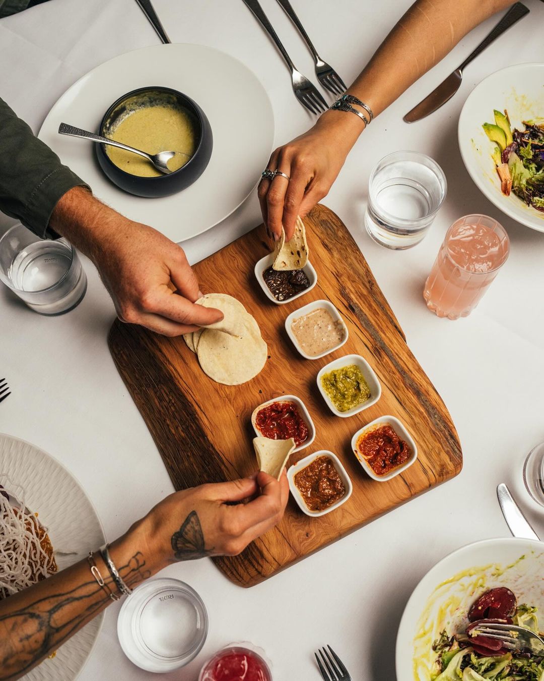 Overhead shot of two people preparing small tortillas with an assortment of sauces on a wooden board, with drinks and salads.