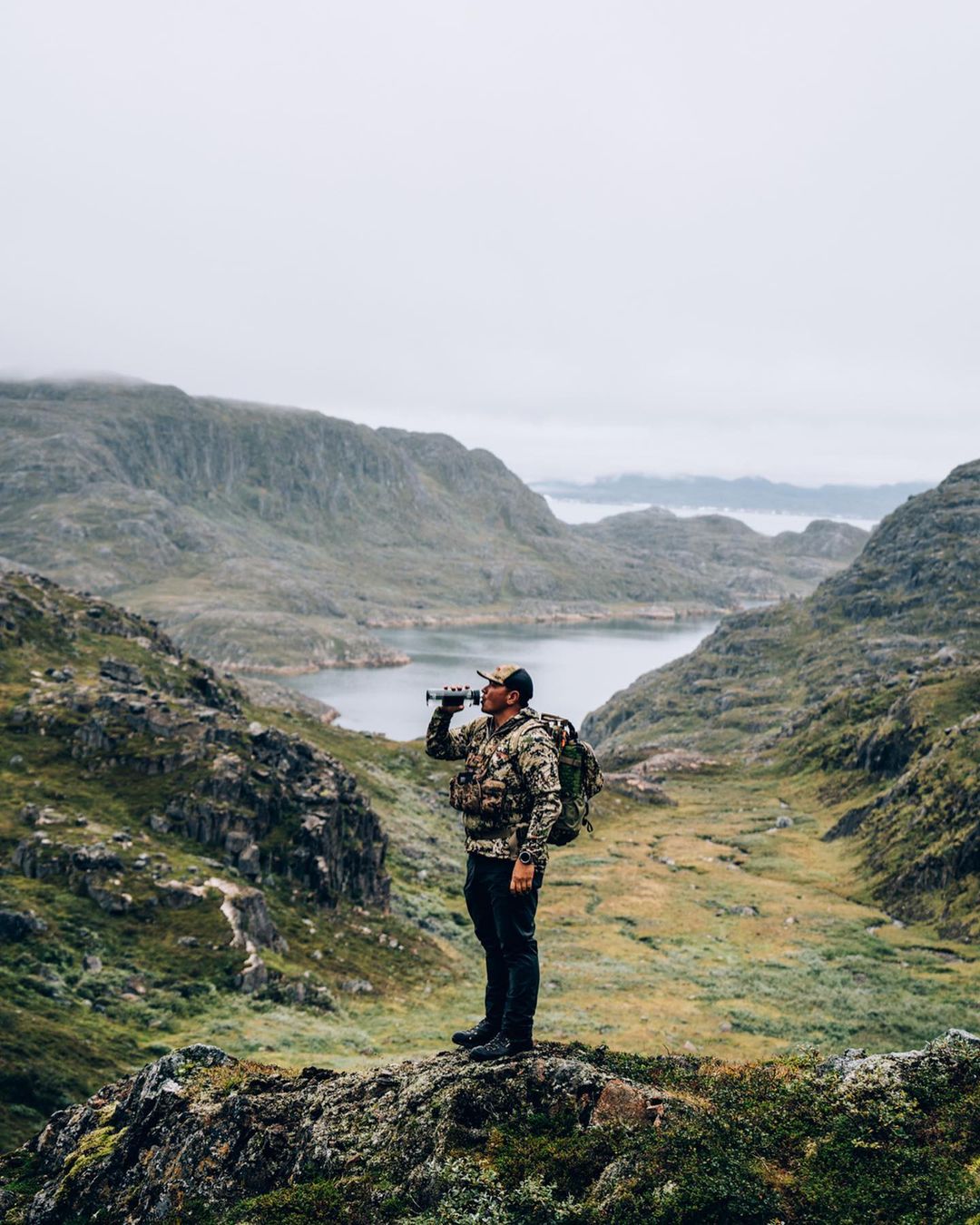 Man in camouflage jacket standing on a rocky overlook drinking from a water bottle, with mountains and a lake in the background.