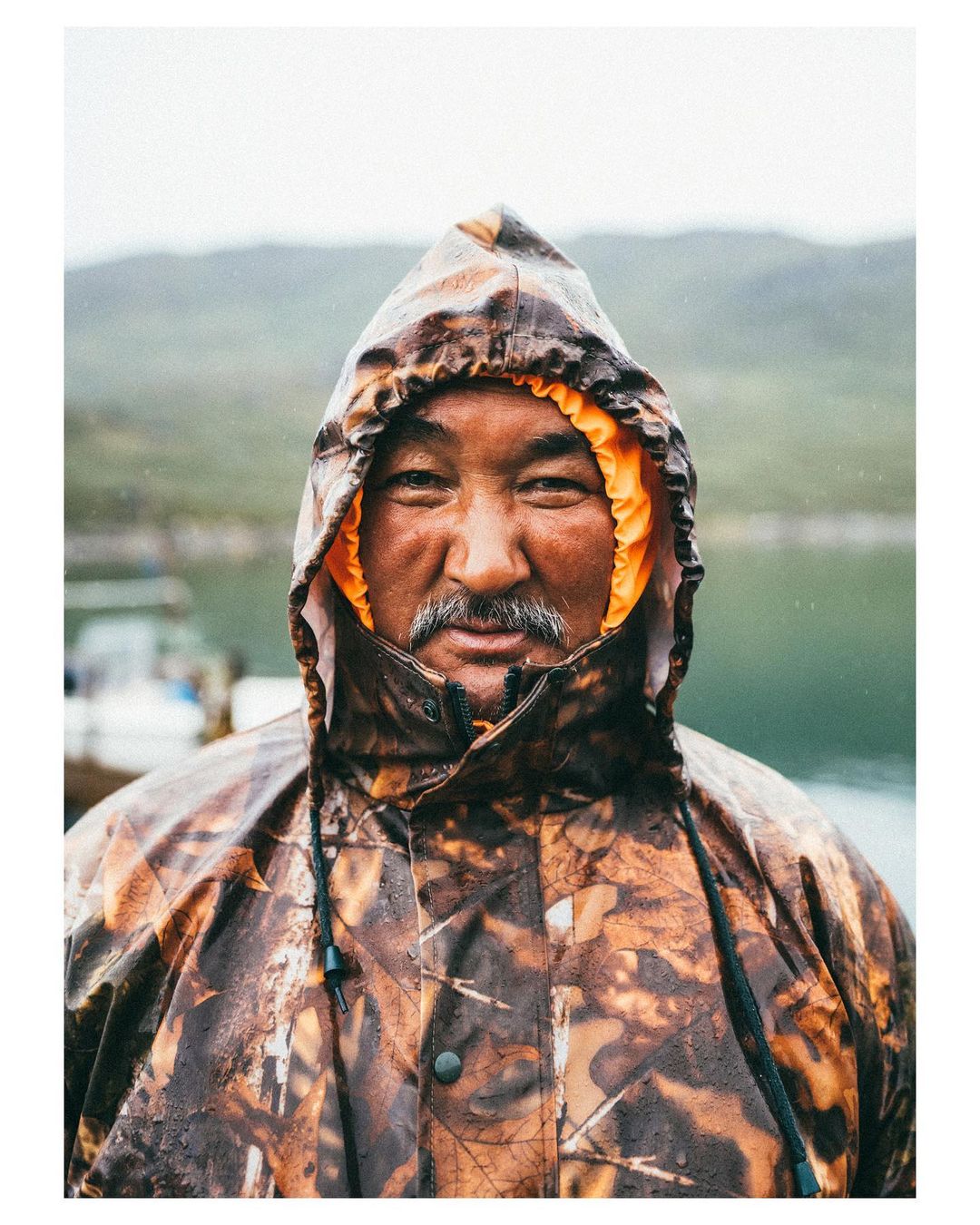 Portrait of a middle-aged man with a mustache wearing a hooded camouflage rain jacket in a rural outdoor setting with blurred background.