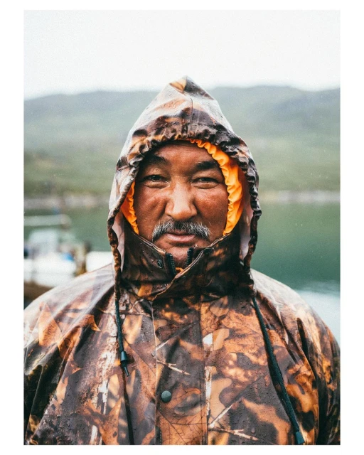 Portrait of a middle-aged man with a mustache wearing a hooded camouflage rain jacket in a rural outdoor setting with blurred background.