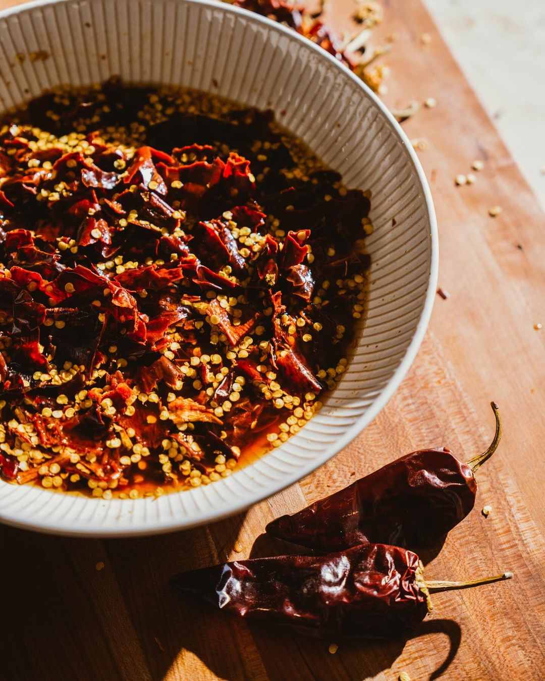 Overhead shot of a white bowl with red chili flakes and seeds, along with two dried chili peppers on a wood surface.